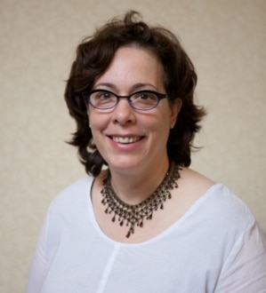 A woman with short brown hair and glasses is smiling at the camera. She is wearing a white top and a large, detailed necklace. The background is plain and neutral.