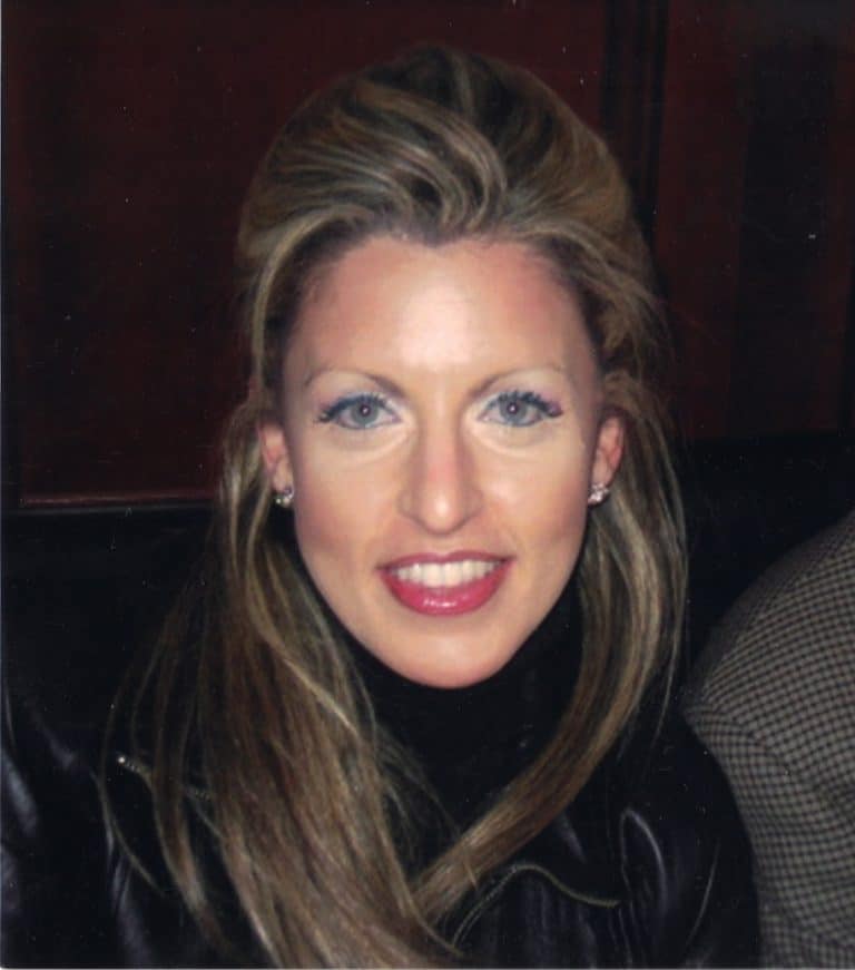 A woman with long blonde hair, light-colored eyes, and light pink lipstick smiles at the camera. She is wearing a black turtleneck and a dark leather jacket. The background is dark and slightly blurred.