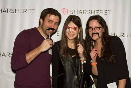 Three people stand in front of a step-and-repeat backdrop with SHARSHERET printed on it, smiling and holding playful props like fake mustaches and glasses on sticks.