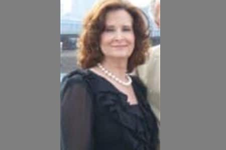A woman with shoulder-length curly brown hair is wearing a black dress with ruffle details and a pearl necklace. She is standing outdoors and smiling at the camera. Part of another person is visible beside her.