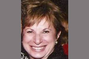 A woman with short light brown hair is smiling widely at the camera. She is wearing a dark top and small earrings. The background is out of focus and the image has gray bars on both sides.