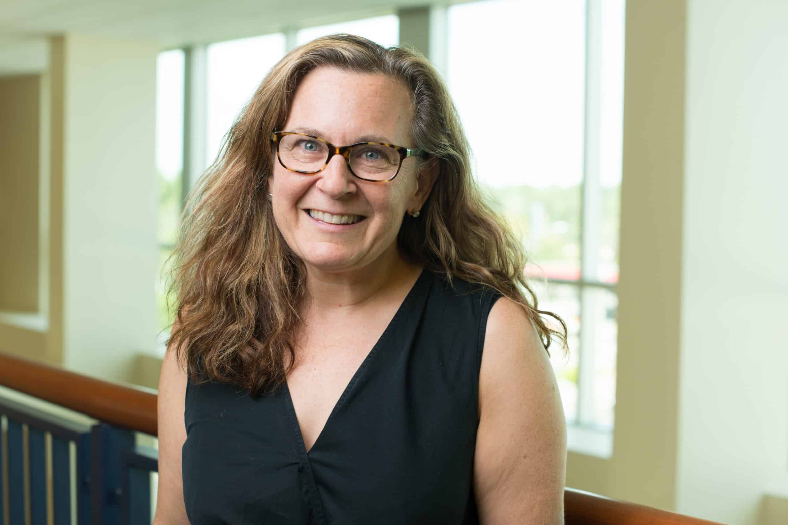 A woman with wavy brown hair and glasses is standing indoors, smiling at the camera. She is wearing a sleeveless black top, and there are large windows with sunlight in the background.