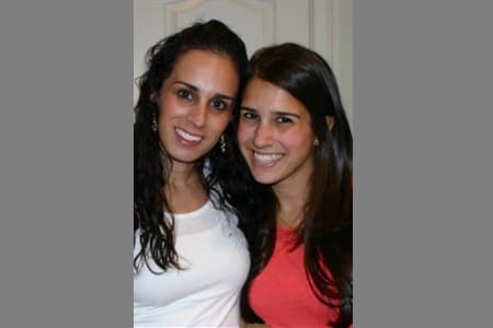 Two women are standing close together indoors, smiling at the camera. One is wearing a white top and has curly dark hair; the other is wearing a red top and has straight dark hair. The background is neutral.