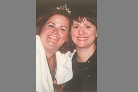 Two women smiling, one wearing a tiara and a white garment draped over her shoulder, while the other wears a dark outfit. They are standing closely together against a dark background.