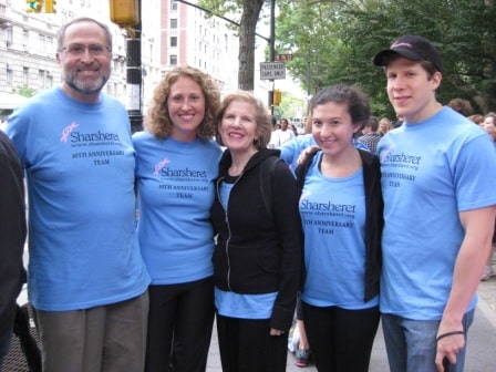 Five people stand outdoors in a row, wearing matching light blue Sharsheret 10th Anniversary Team shirts. Trees and city buildings are visible in the background. They are smiling at the camera.
