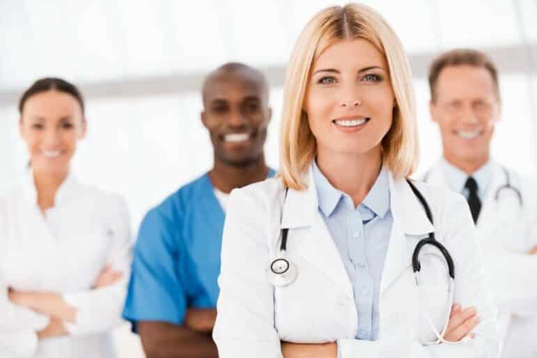 A group of four healthcare professionals, including two women and two men, stand together and smile. The woman in front wears a white coat and stethoscope; the others are in medical attire in the background.