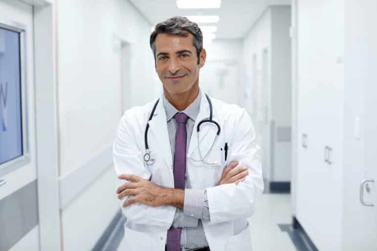 A male doctor with dark hair, wearing a white coat, shirt, tie, and stethoscope, stands in a brightly lit hallway with his arms crossed, looking at the camera and smiling.