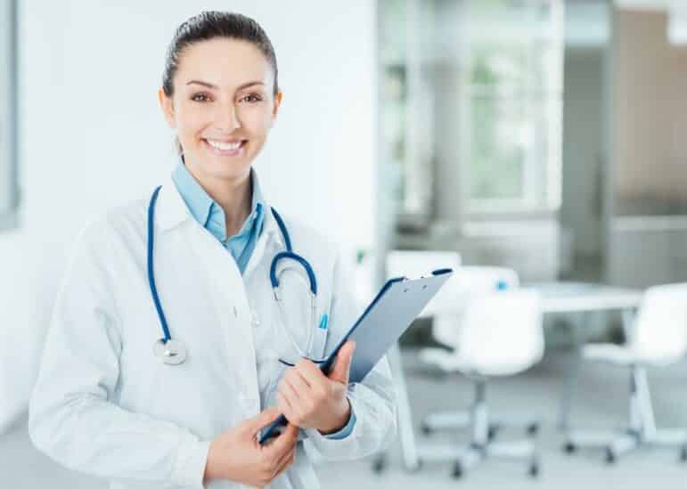 A doctor wearing a white coat and stethoscope stands in a bright, modern medical office, smiling and holding a clipboard.