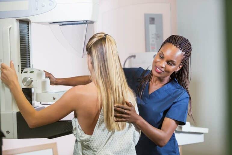 A healthcare professional in navy scrubs assists a woman undergoing a mammogram. The woman stands facing the mammography machine with her side positioned for the scan, wearing a medical gown.