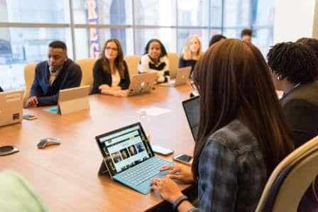 A group of people sit around a large conference table in a modern office, using laptops and tablets, engaged in a meeting or discussion.