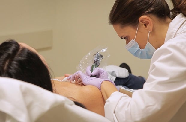 A healthcare professional wearing gloves and a mask performs a medical procedure on a patient lying on an examination table. The professional uses a needle or syringe on the patient’s chest area.