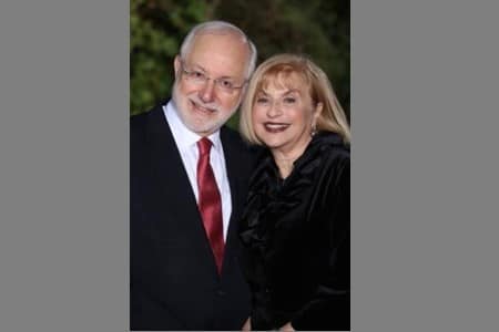 An older man in a suit and red tie stands next to an older woman in a black outfit. Both are smiling and posing together outdoors, with greenery in the blurred background.