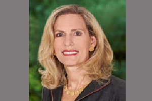 A woman with shoulder-length blonde hair, wearing a dark blazer, gold earrings, and a gold necklace, poses in front of a blurred green background.