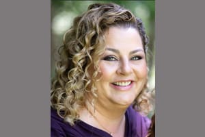A woman with light skin and curly, blonde hair smiles at the camera. She is wearing a purple top and has natural makeup. The background is outdoors and blurred with green tones.