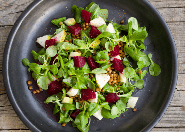 A black plate holds a salad made of leafy greens, diced beets, small cubes of pear, and sprinkled lentils, set on a wooden surface.