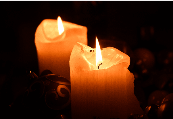 Two lit candles with melted edges burn in a dark environment. The warm orange glow from the flames illuminates their surfaces, and a decorative ornament is partially visible near the base of the candles.