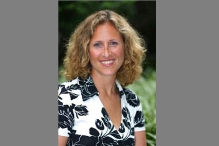 A woman with curly blonde hair, wearing a black and white patterned blouse, is smiling at the camera outdoors with greenery in the background.