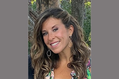 A woman with long wavy brown hair and dangling earrings smiles at the camera. She is outdoors, standing in front of trees, and is wearing a patterned top with a deep neckline.