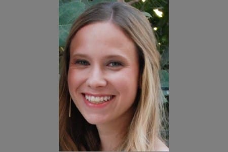 A woman with long, straight blonde hair is smiling at the camera. She is outdoors with green leaves in the background. She is wearing a light-colored top and a single earring is visible.