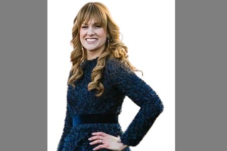 A woman with long, wavy light brown hair stands smiling, wearing a dark textured dress with long sleeves and a black belt, posing with one hand on her hip against a white background.