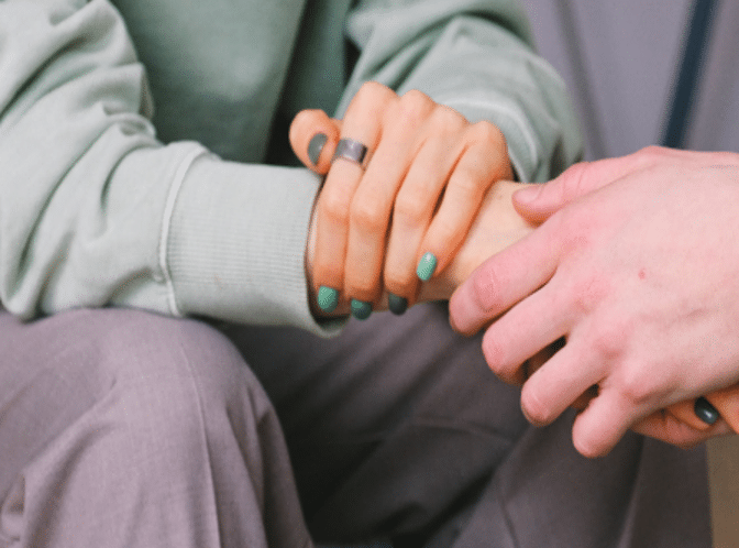 Two women sit at a table having a serious conversation about Jewish breast cancer support. One gestures with her hand while the other listens. Papers, a notebook, and a mug are on the table in front of them.