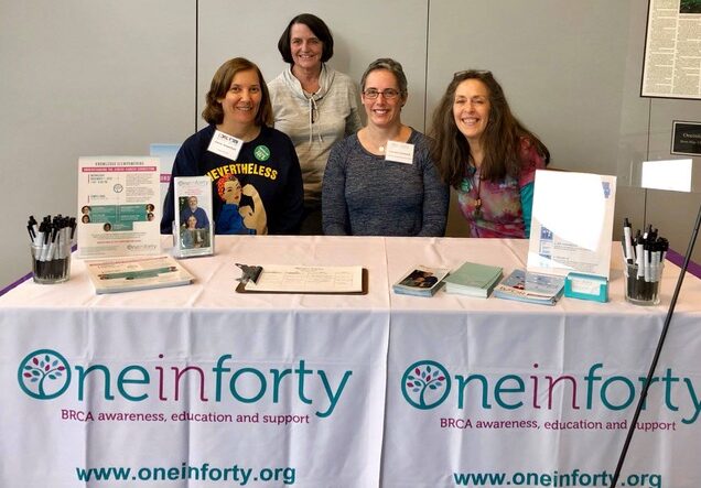 Four women stand and sit behind a table covered with oneinforty banners. The table displays brochures, pens, and information materials about BRCA awareness, education, and support.