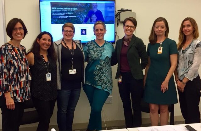 Seven women stand side by side in a meeting room, smiling at the camera. Behind them, a computer screen displays a website with text and images. Some women wear ID badges, and a table is partially visible in front.