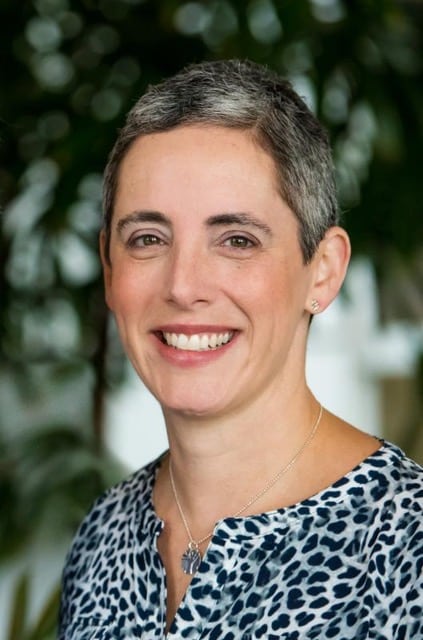 A person with short gray hair and light skin is smiling at the camera. They are wearing a blue and white patterned top and a silver necklace. The background is blurred with greenery visible.