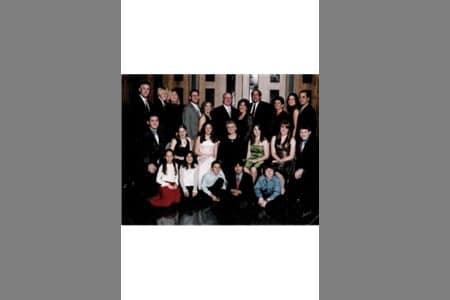 A group of people of various ages, dressed in formal attire, pose together indoors in several rows, with adults standing in the back and children sitting or kneeling in front.