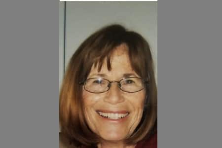 A middle-aged woman with straight, shoulder-length brown hair and glasses smiles at the camera. She is standing in front of a neutral background.
