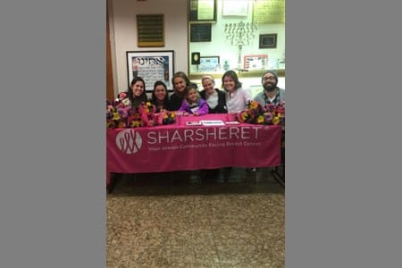 Seven people sit behind a table covered with bouquets of flowers and a pink tablecloth displaying the Sharsheret logo and tagline about breast cancer support. Plaques and flyers are visible on the wall behind them.