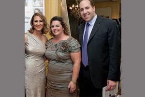 Three adults pose together indoors at a formal event. Two women stand on the left wearing metallic dresses, and a man stands on the right in a dark suit with a purple tie. They are smiling at the camera.