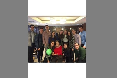 A group of thirteen people pose indoors, some standing and some sitting. One person in the front row holds a green ball. They are in a well-lit room with a modern ceiling light visible above them.