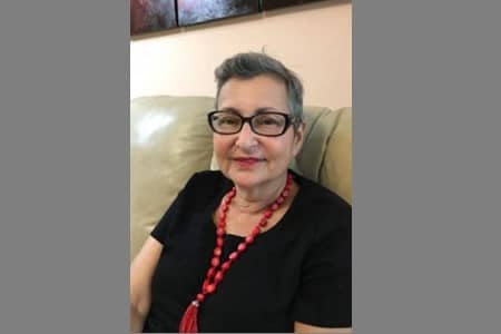 An older woman with short gray hair and glasses sits on a beige couch, wearing a black top and a long red beaded necklace. She is looking at the camera with a slight smile.