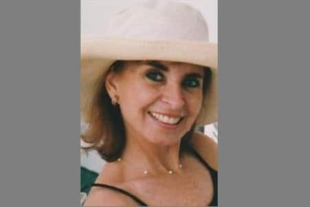 A woman wearing a wide-brimmed light-colored hat and a black top smiles at the camera. She has shoulder-length brown hair and is wearing earrings and a necklace. The background is plain and light-colored.