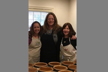 Three women stand side by side indoors, smiling at the camera. They are wearing aprons and standing behind a table filled with several pumpkin pies. A white window with blinds is in the background.