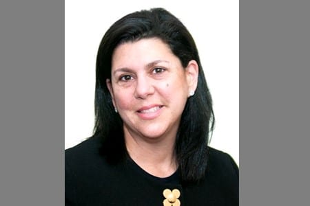 A woman with straight, shoulder-length black hair is smiling slightly. She is wearing a black top with a decorative gold detail near the neckline. The background is plain and light-colored.
