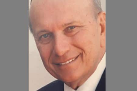 A close-up photo of an older man with light skin, short gray hair, and blue eyes, smiling and wearing a dark jacket and white shirt. The background is plain and light-colored.
