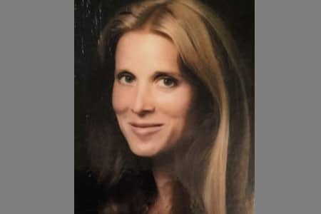 A woman with long, straight, light brown hair is smiling softly at the camera against a dark background. She is facing slightly to the left.