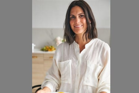 A woman with shoulder-length dark hair, wearing a white button-up shirt, stands in a kitchen and smiles at the camera. The background shows a light-colored countertop with fruit and kitchen items.