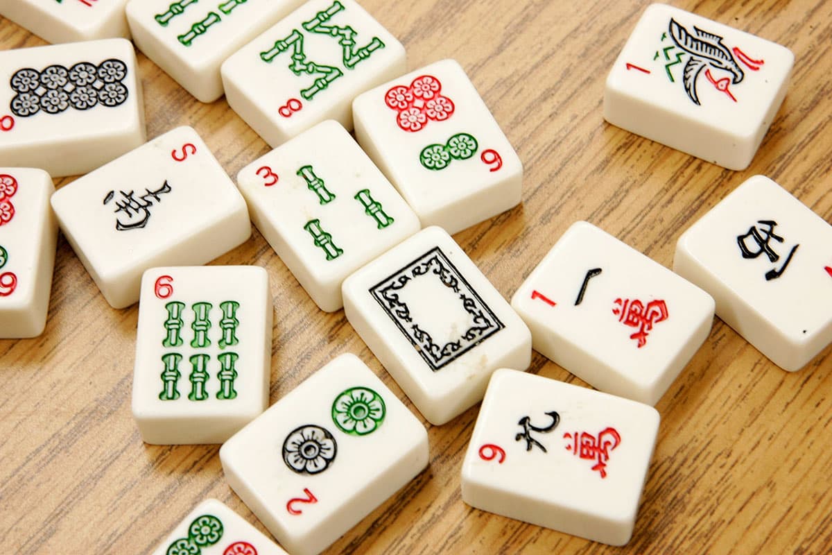 Mahjong tiles with various symbols, numbers, and colors are scattered on a wooden surface. The tiles include circles, bamboo sticks, characters, and a bird illustration.
