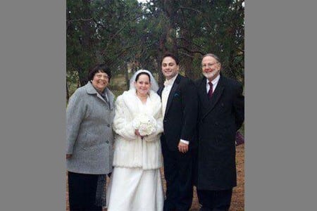 Four adults pose outdoors for a photo. The two people in the middle wear wedding attire, with the bride in a white dress and fur coat, holding a bouquet. The other two wear dark overcoats. Trees and grass are in the background.