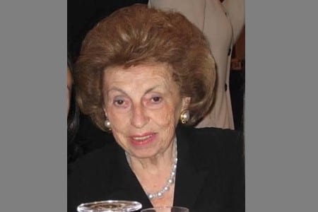 An older woman with styled brown hair, wearing pearl earrings, a pearl necklace, and a black top, is seated indoors with a glass in front of her. The background is blurred and partially obscured.