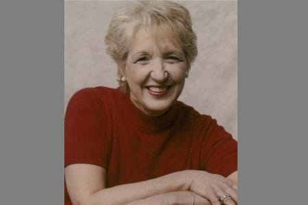 An older woman with short, light-colored hair smiles at the camera. She is wearing a red short-sleeved top and small earrings, with her arms resting in front of her against a light, neutral background.