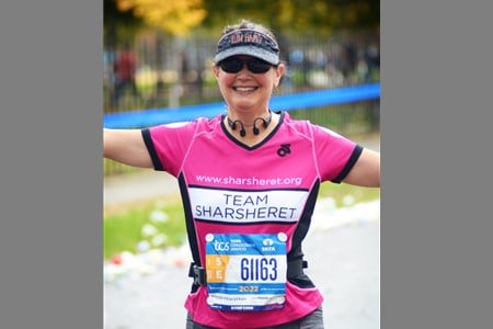 A smiling runner wearing a pink Team Sharsheret shirt, sunglasses, and a race bib stands with arms outstretched. The race appears to be outdoors with blurred trees and a fence in the background.