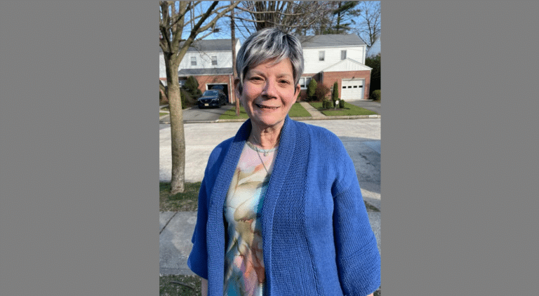 An older woman with short gray hair stands outside on a residential street. She is wearing a blue sweater over a patterned top, and there are houses, cars, and leafless trees in the background.