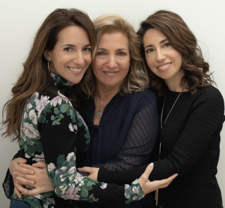Three people, two with dark brown hair and one in the middle with lighter blonde hair stand in front of a white background holding each other close.