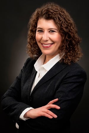 A dark haired person wearing a black sweater and white shirt in front of a black background.