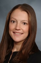 A woman with straight, long brown hair is smiling at the camera. She is wearing a black top and is posed against a plain, gray background.