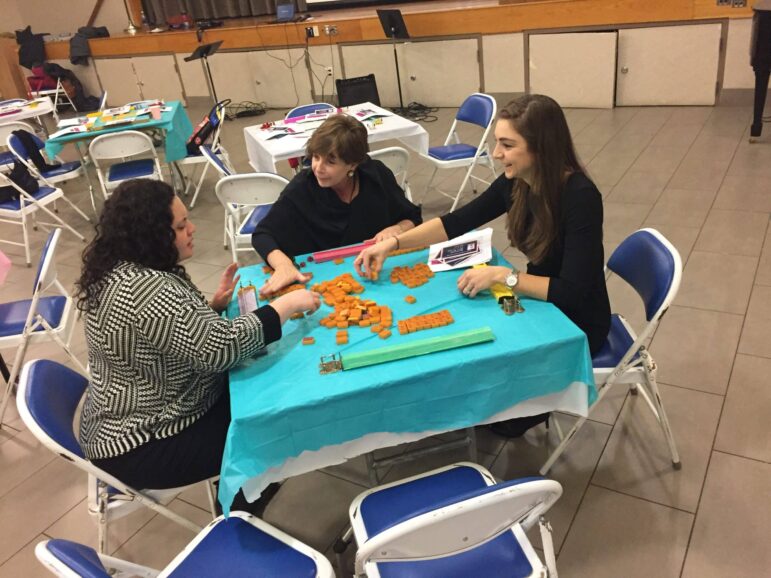 Three women sit around a table covered with a blue tablecloth, arranging orange counting blocks. Papers, folders, and other educational materials are scattered on the table. Empty chairs and tables are in the background.